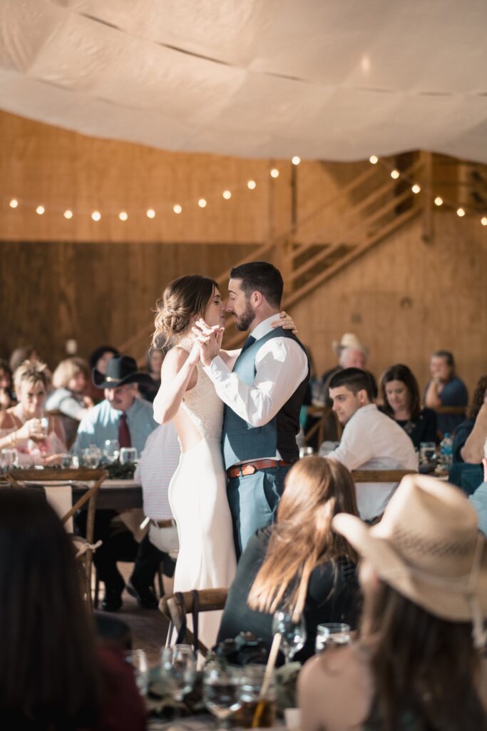 bride and groom's first dance in the barn at Diamond Cross ranch in front of all of their wedding guests with lights hanging and twinkling above