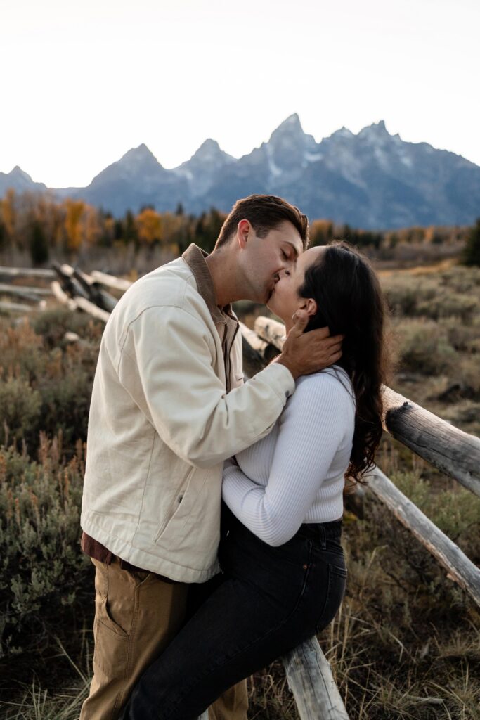 Newly engaged couple embrace and kiss one another as the sun sets below the Tetons of Jackson Hole Wyoming on their fall surprise engagement session