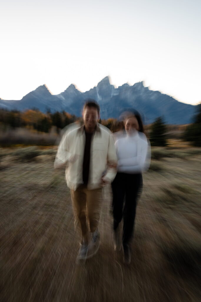 engaged couple run towards the camera as the background and the view of the Tetons in Jackson Hole blurs in the background at sunset 
